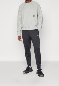 Grauer Baumwollpullover mit Rundhalsausschnitt und schwarzem Logo-Patch, kombiniert mit dunkelgrauen Jogginghosen, die ein weißes Nike-Logo aufweisen. Schwarze Sneakers.