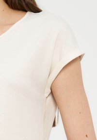 Κοντινό πλάνο ενός ατόμου που φοράει μπλούζα κοντομάνικη, σε ανοιχτό μπεζ χρώμα, με γυρισμένη μανσέτα στο μανίκι.