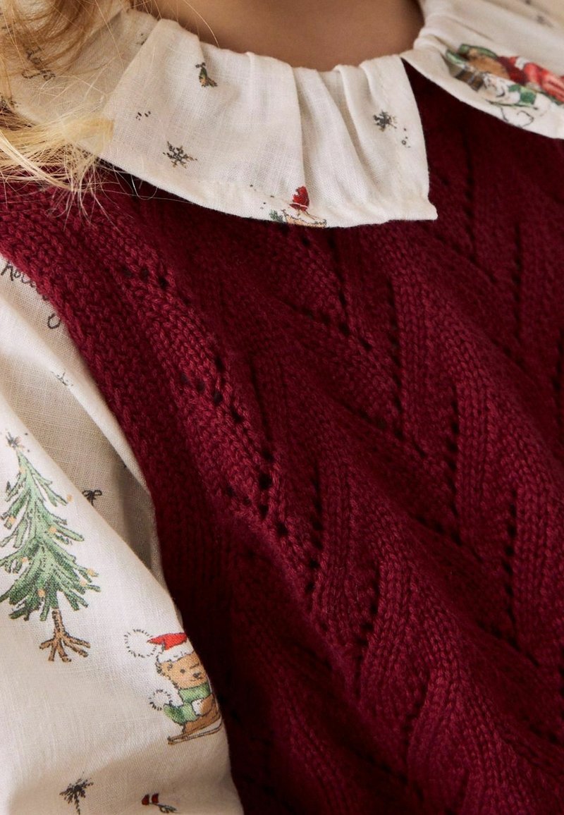 Gros plan d’une personne portant un pull en maille bordeaux sur un chemisier blanc avec un petit motif sapin de Noël et ours sur la manche et le col.