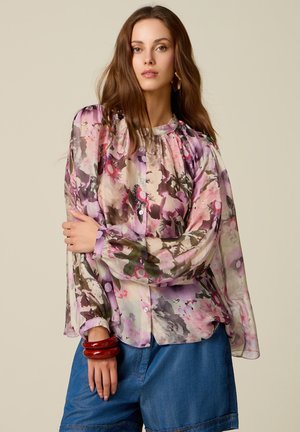 Frau mit langen braunen Haaren, die eine transparente, florale Bluse mit Puffärmeln und lockere blaue Jeansshorts trägt, ergänzt durch auffällige rote Armbänder.