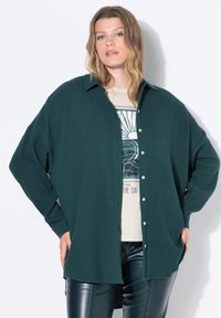 Camicia a maniche lunghe verde scuro con un tessuto texture, abbottonata sul fronte e dalla vestibilità oversize. Indossata sopra una t-shirt beige con grafica e pantaloni in pelle.