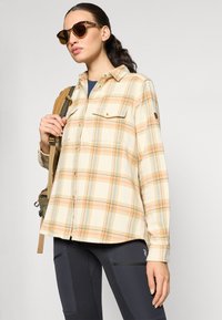 Camicia di flanella a quadri leggeri in tonalità beige, arancione e blu; presenta due tasche sul petto, chiusura con bottoni e orlo curvo. Leggings neri in vista.