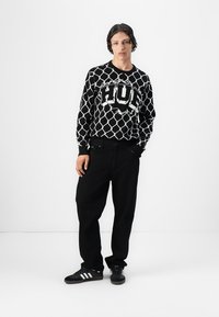 Sorte og hvide mønstrede sweater med stort "HUG" logo, parret med sorte bukser og sorte sneakers med hvide striber. Glat tekstur.