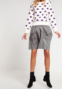 Sweatshirt blanc avec motif de logo rouge et bleu, accompagné de shorts gris plissés. Des bottes noires à la cheville complètent la tenue.