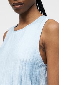 Κοντινό πλάνο ενός ατόμου που φοράει ανοιχτό μπλε, αμάνικο, υφασμάτινο πάνω μέρος με πλεγμένα μαλλιά σε απλό φόντο.