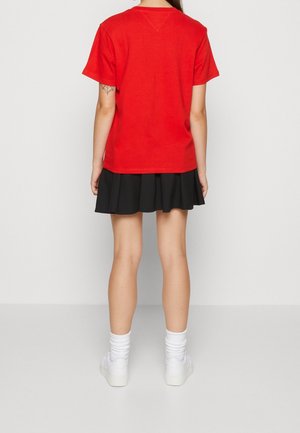 Persoon die met de rug naar de kijker staat, gekleed in een rood T-shirt, zwarte geplooide rok, witte sokken en witte sportschoenen tegen een effen achtergrond.