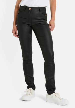 Pantalon en simili cuir noir à coupe slim, avec poches avant et studs discrets sur les côtés. Associé à des baskets blanches à lacets.