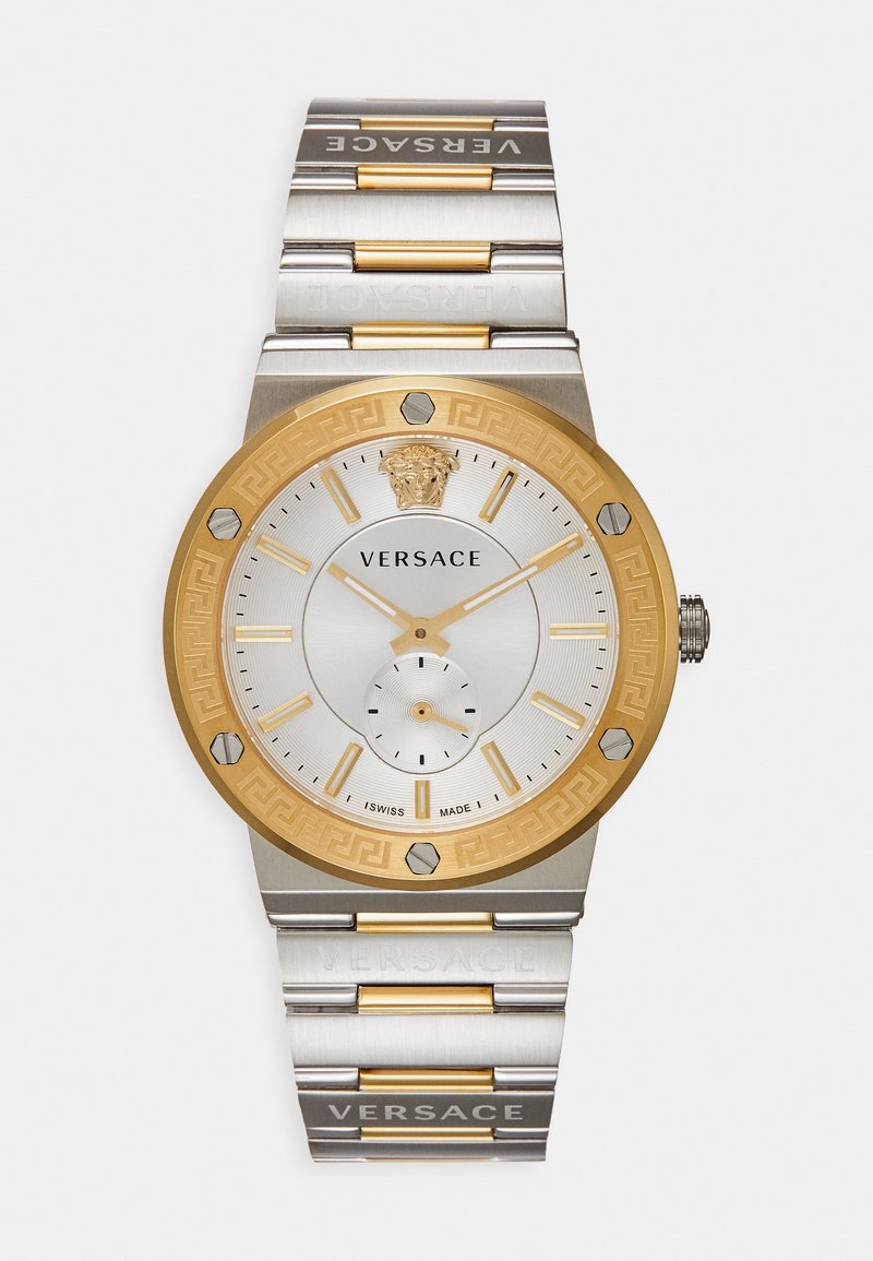 Versace kell, millel on hõbedane alused, kuldsetooniline äär ja kuusnurkse kujuga aktsendid. Roostevabast terasest käevõru kuldsete detailidega. Šveitsi valmistatud.