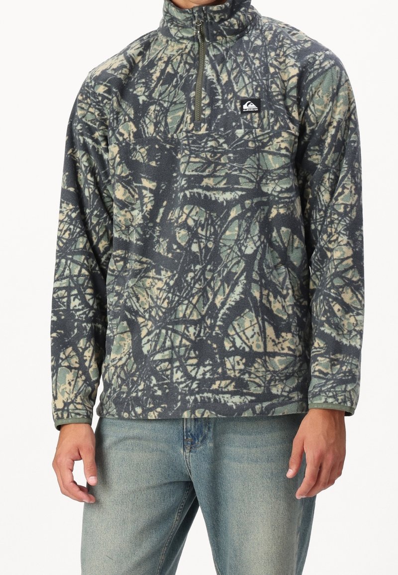 Fleece-pooler med høj krave, halv lynlås lukning, i camouflage mønster af grønne og brune nuancer, med et logo-patch på den øverste bryst.
