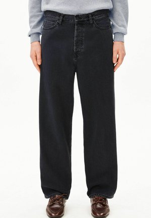 Jean noir droit porté avec un sweat gris et des chaussures marron à lacets, visible de la taille aux pieds.