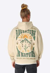 Cremefarbener Kapuzenpullover mit einem großen, bunten Grafikelement auf dem Rücken, das Berge, Sonne und dekorative Elemente zeigt. Text: "ABENTEUER IN DER NATUR."