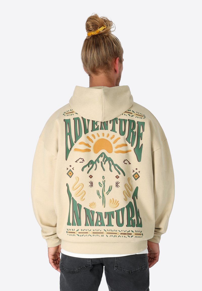 Cremefarbener Kapuzenpullover mit einem großen, bunten Grafikelement auf dem Rücken, das Berge, Sonne und dekorative Elemente zeigt. Text: "ABENTEUER IN DER NATUR."
