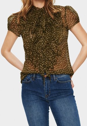 Mujer con blusa transparente verde oliva con lunares y volantes combinada con vaqueros azules, manos apoyadas en las caderas.