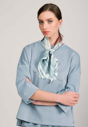 Joven con sudadera azul claro y pañuelo de seda estampado atado al cuello, brazos cruzados, fondo neutro.