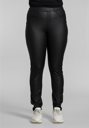 Person, die schwarze Kunstlederhosen und weiße Turnschuhe trägt und vor einem neutralen grauen Hintergrund steht.