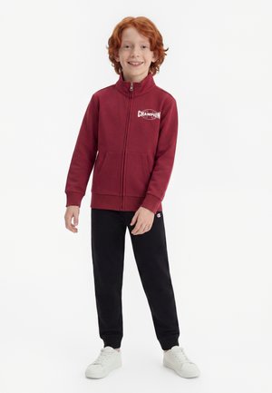 Smilende rødhåret dreng iført en bordeaux Champion zip-up sweatshirt, sorte joggingbukser og hvide sneakers, stående på en hvid baggrund.
