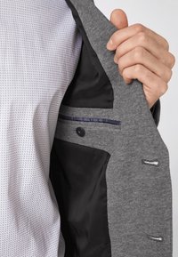 Giacca grigia con un esterno testurizzato, foderatura in satin nero, tasca interna con bordatura blu navy e chiusura a bottone. Camicia sotto a motivi ben visibili.