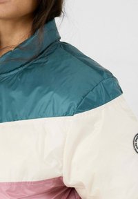 Persona che indossa una giacca bloccata nei colori teal, bianco e rosa con una toppa sulla manica, su uno sfondo chiaro e semplice.