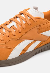 Orange sportssko med ovandel i mocka, vita läderdetaljer, perforerade inslag och en texturerad brun gummisula.