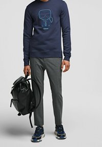 Man som bär en marinblå sweatshirt med grafik, grå byxor, blå sneakers och håller en svart läderryggsäck i ett av remmarna.