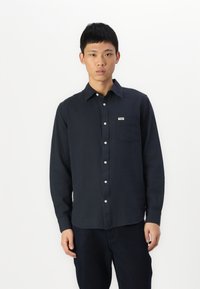 Camicia button-up a maniche lunghe di colore navy realizzata in cotone. Presenta una tasca, un colletto a punta e bottoni bianchi. Texture liscia con design minimale.