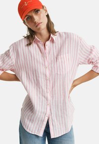 Camicia button-up a righe rosa e bianche con taschino sul petto, realizzata in tessuto leggero. Abbinata a un cappellino arancione a contrasto.