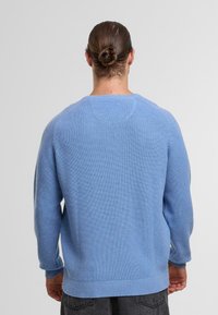 Person mit braunen Haaren zu einem Dutt gebunden, trägt einen hellblauen Strickpullover und dunkle graue Hosen, von hinten vor einem schlichten Hintergrund dargestellt.