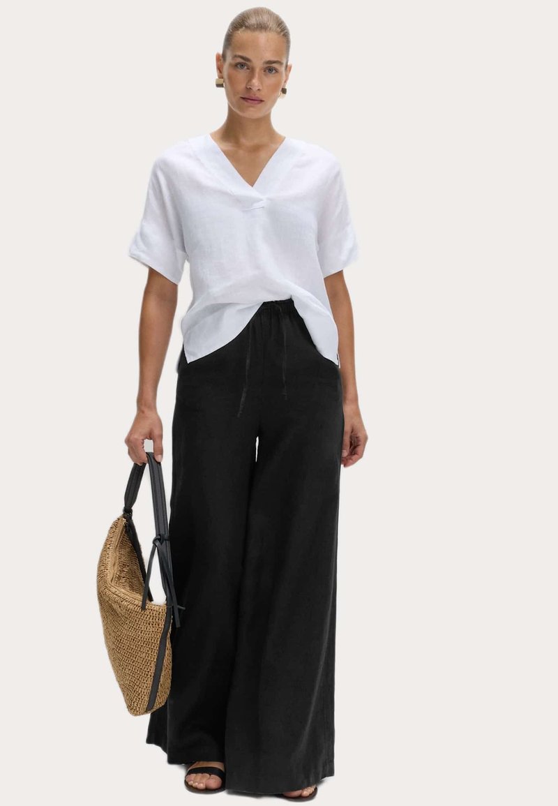 Biała lniana bluzka z krótkim rękawem i dekoltem w kształcie litery V, zestawiona z czarnymi spodniami o szerokich nogawkach. Uzupełniona plecioną torbą i czarnymi sandałami na paski.