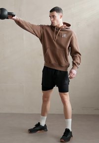Barna pamut kapucnis pulóver elülső zsebből, fekete rövidnadrággal és fekete sportcipővel párosítva. A téma egy kézzel kettlebellt tart.