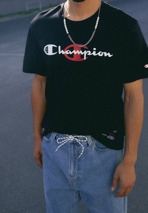 Man draagt een zwart Champion T-shirt en lichtblauwe spijkerbroek met een wit-zwarte trekkoord, staand in de buitenlucht.