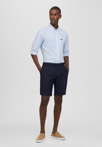 Camicia button-up a righe azzurre con maniche arrotolate e pantaloni corti blu navy. Scarpe slip-on beige completano l'outfit. Design semplice, stile casual.