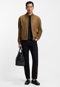 Brun bomberjacka i mocka, svart T-shirt, svarta byxor och svarta loafers. Håller en svart tygväska med struktur. Enkel, modern design.