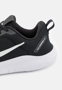 Sort sportssko med tekstureret mesh-overside, hvide accenter og en stor bageste rem. Har en fleksibel sål og mærket "Flex Experience 12".