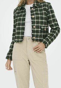 Femme portant une veste en tweed à carreaux verts et blancs avec des boutons dorés, par-dessus une chemise blanche et un pantalon en velours côtelé beige avec de grandes poches.