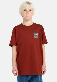 Camiseta de algodón roja con un escote redondo, mangas cortas y un parche de tela con gráficos de olas en el lado izquierdo del pecho.