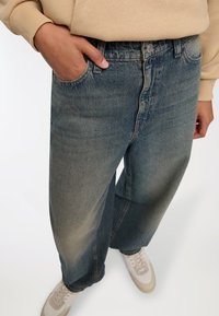 Jeans in denim oversize blu chiaro con lavaggio sbiadito, cinque tasche e chiusura con bottone, abbinati a sneakers bianche.