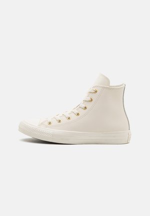 Højtliggende sneakers i off-white læder med guldøjer, tekstureret gummisål og rundet tå; slankt design med minimale detaljer.