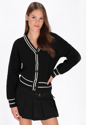 Junge Frau trägt eine schwarze, geknöpfte Strickjacke mit weißen Streifen und einen schwarzen Faltenrock, posiert mit einer Hand in der Hüfte.