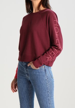 Kvinde iført en kort burgunderrød sweatshirt med "HOLLISTER" trykt på ærmet og blå højttaljede jeans, hånd i lommen.