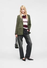 Žena v pruhovanom svetri, olivovom blejzeri, čiernych džínsach a podpätkoch, ktorá drží čiernu kabelku, stojí pred jednoduchým pozadím.