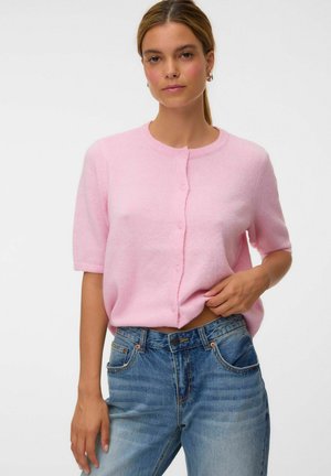 Femme portant un pull rose clair à manches courtes boutonné et un jean bleu taille haute, debout devant un fond blanc uni.