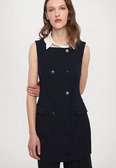 Vestă fără mâneci de culoare bleumarin cu design dublu, nasturi negri, două buzunare frontale și guler cu poale, purtată peste o cămașă albă.