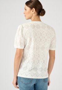 Witte kanten top met korte pofmouwen, met een diamanten patroon en open geweven textuur. G gedragen met blauwe spijkerbroek.