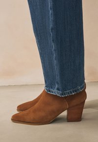Bottes cavalières en daim marron avec un bout pointu et un talon empilé texturé, associées à un jean droit bleu clair légèrement déchiré.