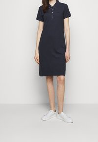 Námořnická modrá polo šaty vyrobené z bavlny, s límečkem, třemi knoflíky, krátkými rukávy a rovný střih. Nosené s bílými teniskami.