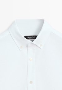 Weißes Baumwollhemd mit klassischem Kragen und einer Leiste mit drei Knöpfen. Es zeichnet sich durch eine dezente Struktur und ein Etikett mit Marken- und Herkunftsangabe aus.