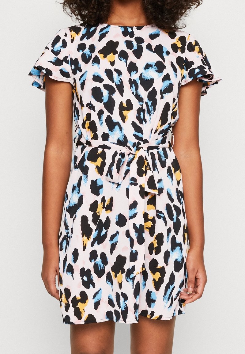 Femme portant une robe à manches courtes avec une base rose et des taches abstraites de léopard noires, bleues et jaunes, nouée avec une ceinture assortie à la taille.
