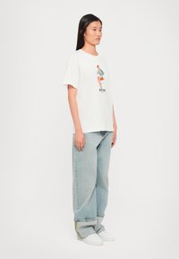 T-shirt en coton blanc avec un imprimé graphique coloré et une coupe décontractée, associé à un jean en denim bleu clair et des baskets blanches.