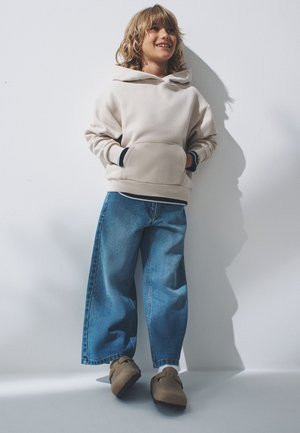 Niño con el pelo hasta los hombros apoyado en una pared, lleva una sudadera beige con capucha, vaqueros anchos azules y zapatos marrones de tipo slip-on, sonriente y mirando hacia arriba.