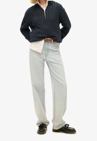 Marineblauer Pullover mit Kabelstrickmuster und halbem Reißverschluss, kombiniert mit hellblauen Jeans mit geradem Bein und schwarzen Schnürschuhen. Akzent durch braunen geflochtenen Gürtel.